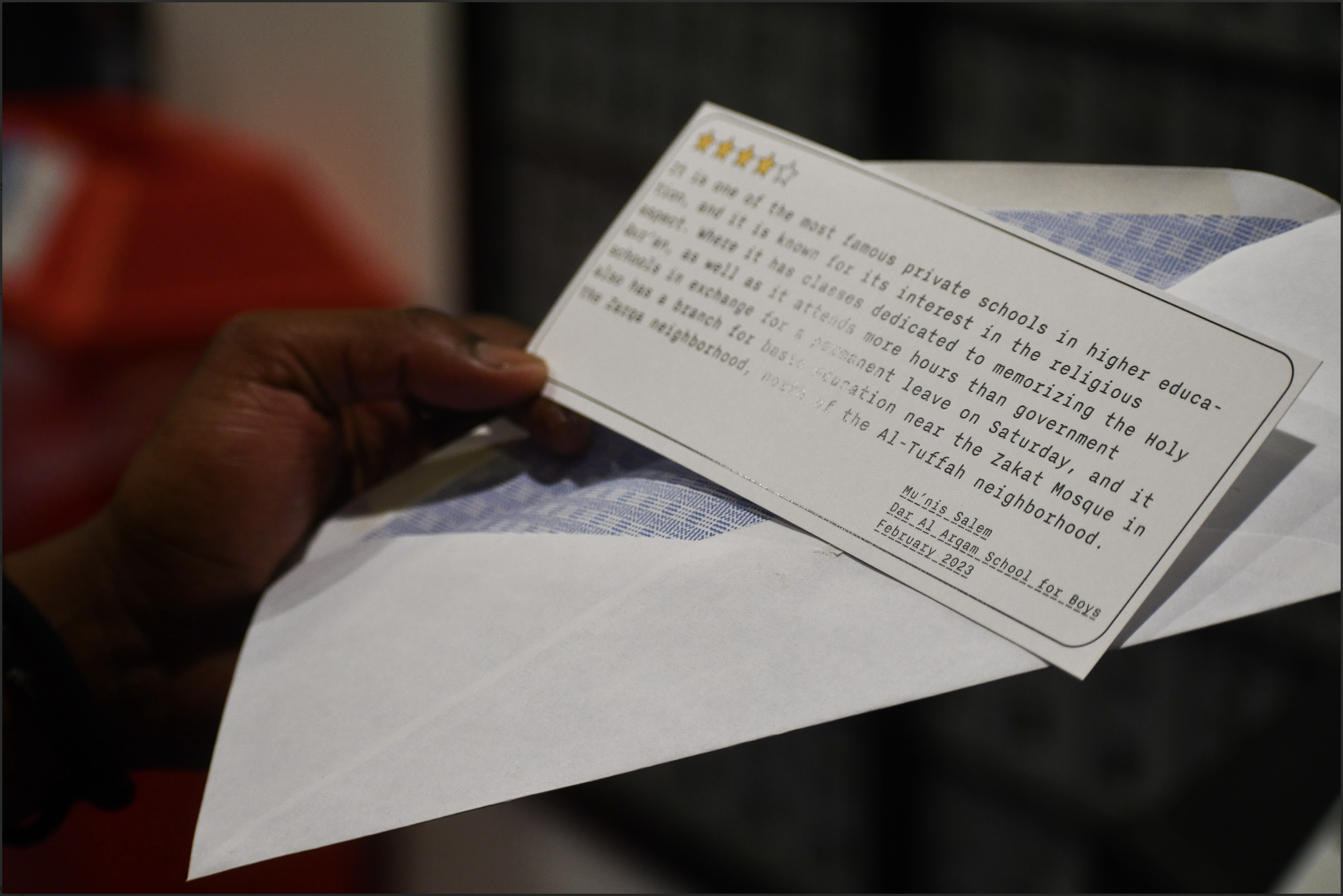 A hand holding a postcard-style review of a Gazan school, pulled from an envelope